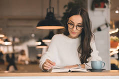 有吸引力的年轻妇女在眼镜读书书在桌与咖啡杯子在咖啡馆