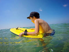 快乐男孩在盖帽在游泳树干站立在海滩在金黄沙子并且举行一个游泳板, 黄色蓝色的摇摆的板材对蓝天和海以小波浪在阳光下.