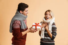 一对穿着毛衣和围巾的年轻夫妇的肖像, 站在米色的背水一战上, 快乐的男人送给他的女朋友一份礼物