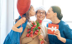 母亲节快乐！孩子的女儿在祝贺妈妈和奶奶送给他们的花郁金香。奶奶，妈妈和女孩笑着和拥抱。家庭假日和团聚.
