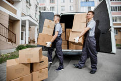 两个穿着制服的年轻英俊微笑的工人正在卸货, 面包车里装满了箱子。公寓楼在背景中。房子移动, 搬运工服务
