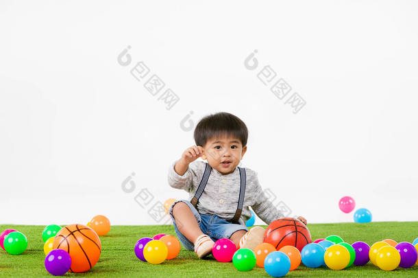 工作室的肖像可爱的, 亚洲幼儿男孩穿着牛仔工作服, 长袖 t恤, 看着他的手, 坐在人造绿草与五颜六色的球, 在孤立的白色背景