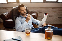 年轻的英俊商人坐在椅子上, 在自己的办公室里看着雪茄。他把腿放在桌子上, 手里拿着日记。威士忌的玻璃和石墨烯.