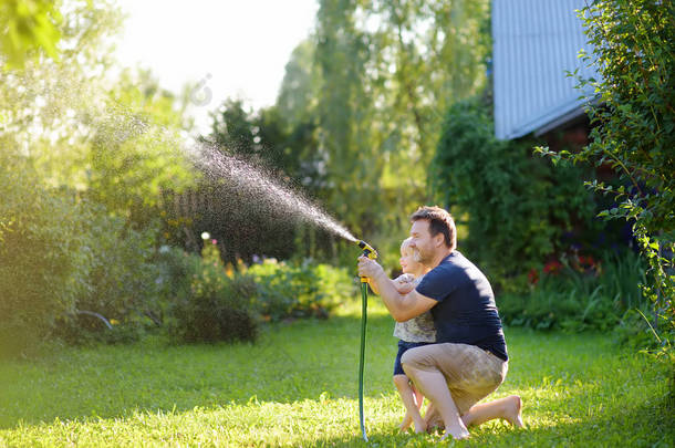 有趣的小男孩与他的父亲玩花园软管在阳光明媚的后院。学龄前儿童在用水喷雾玩乐.