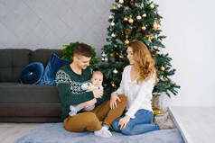 客厅里一个快乐的微笑和欢乐的家庭的肖像，装饰圣诞节。父亲抱着孩子，母亲看着他们，微笑着。家人坐在地板上。舒适的概念，庆祝在Chri新年