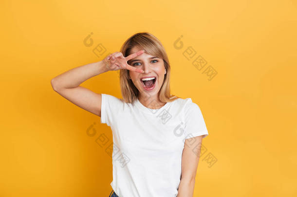 乐观情绪化的年轻漂亮的金发女郎在黄墙背景上摆出孤立，穿着白色休闲T恤，显示和平姿态.