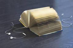 有声读物概念。耳机，在深色木制背景上打开一本书。听有声读物，而不是阅读。耳机线位于页面。在地板上打开书.