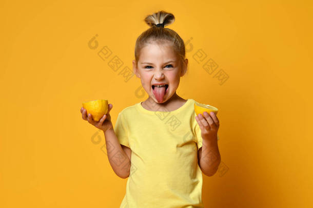 穿着黄色T恤的可爱的小女孩，手里拿着一半新鲜酸成熟的柠檬果，在黄色的背景上露出舌头