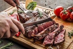 在木制切菜板上手工切碎中等稀罕的烤战斧肉或肋眼牛排，在深色背景下烤蔬菜，关门