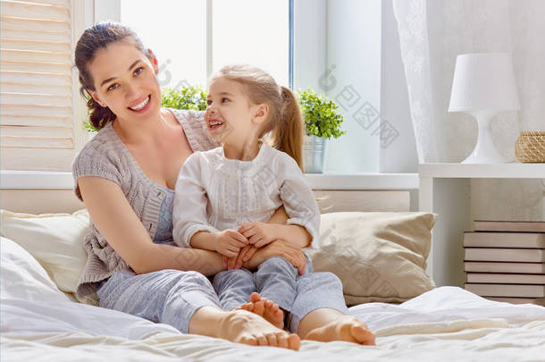 一个漂亮的女孩和她的母亲享受着阳光灿烂的早晨。在家里玩得很开心孩子从睡梦中醒来。家人在卧室的床上玩耍.  