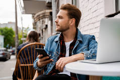 自由撰稿人持有手机，并在室外使用笔记本电脑工作。坐在街上的城市咖啡馆里，独自放松梦想的时光。在线通信和互联网技术概念