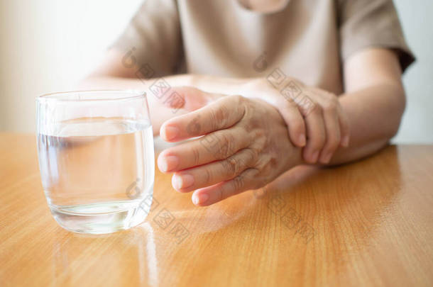 老年妇女的手颤抖着症状伸出来在木桌上取一杯水。握手的原因包括帕金森病、中风或脑损伤。精神健康神经紊乱.