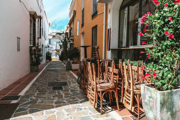 在西班牙，一<strong>大</strong>早就可以看到一家关门的咖啡店。明亮的阳光映衬着堆叠的椅子和五颜六色的黄色和白色的建筑物。美丽而狭窄的西班牙街道，开着红花.