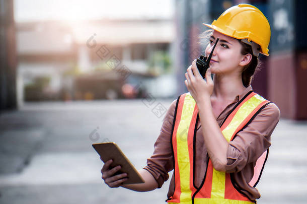 年轻自信的<strong>高加索</strong>女工程师，使用无线电通信或对讲机，头戴黄色安全帽，检查货物货轮装卸集装箱的情况，以便进出口