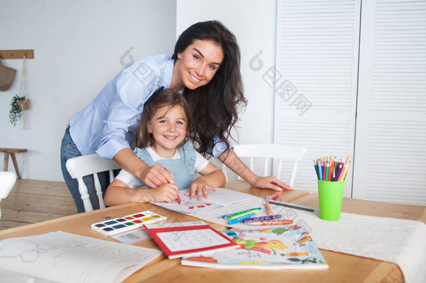 微笑着母亲和女儿准备上课，并用铅笔和颜料在桌边画画。学前教育的父母和学生。秋天的第一天。小班的女生，回学校.