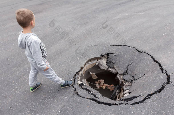 小孩站在沥青路面大坑边.城市街头儿童受伤的危险。道路表面孔