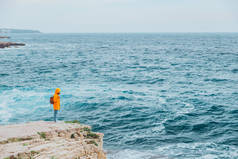 身穿黄色雨衣、背着背包的女人站在悬崖上凝视着励志的大海