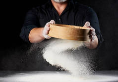 厨师身穿黑色制服的人手里拿着一个圆圆的木制筛子，在黑色的背景上筛出白面粉，颗粒向不同方向飞散