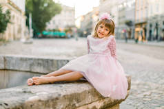 迷人的白种人小女孩穿着时髦的粉色公主礼服，没有鞋子，坐在古城背景街上的古老石泉上的浪漫画像