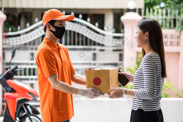 带着面罩和橙色制服手套的快递员，给了带着面罩的亚洲微笑妇女纸箱包裹。网上包装是用摩托车送回家的。COVID-19大流行病。新常态