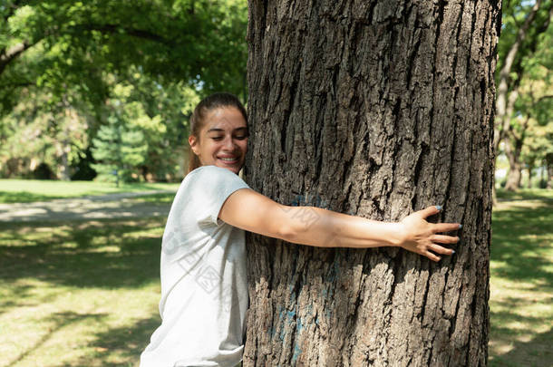 美丽的微笑着的年轻而积极的、快乐的环境主义者女孩拥抱着公园里的大树，热爱大<strong>自然</strong>，想<strong>保护</strong>好环境，<strong>保护</strong>好森林