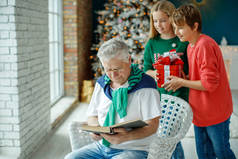 祖父和孙子孙女们在圣诞树下装饰着圣诞节的房间里。圣诞节假期的概念。对比摄影.