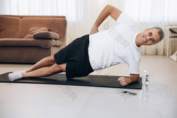 快乐的老年人在瑜伽垫上做侧板运动。如何在检疫观念上保持健康。健康的生活方式运动员训练和看着镜头。背景的现代客厅.