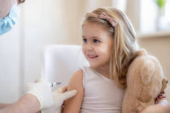 儿童免疫概念。快乐可爱的金发碧眼的小女孩拿着玩具，拿着流感疫苗，不怕针筒针。医生在诊所或诊所给勇敢的儿童注射Covid-19疫苗