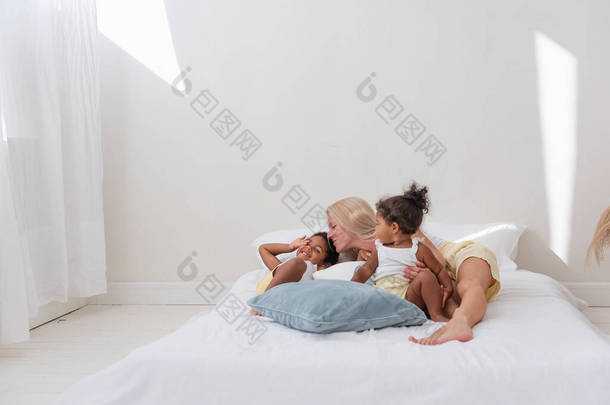 年轻的白人金发母亲玩耍，躺在白色的床上与非洲裔美国人的女儿的儿子。多民族<strong>家</strong>庭的快乐早晨。女人拥抱着,温柔地抚摸着孩子.<strong>简约</strong>的白色内部