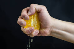 一个人的手握住一个橙子，握紧他的拳头，在黑色的背景下挤压果汁。早餐用新鲜橙汁.
