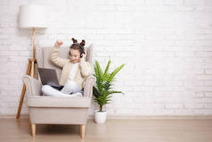人、技术、互联网和在线教育概念- -戴着耳机坐在扶手椅上的可爱少女的肖像，使用笔记本电脑，在白砖墙背景上听音乐，并附有复制空间