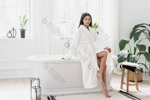 迷人的年轻亚洲女人坐在轻盈宽敞的浴室里，绿色的植物穿着白色浴衣，在享用完早晨的日常活动后露出腿。自理概念