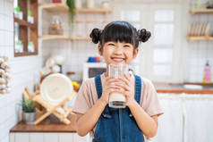 在家里厨房里拿着一杯牛奶的亚洲可爱小孩的画像。学龄前小女孩或女儿笑着呆在家里，开心地喝牛奶，然后看着相机.