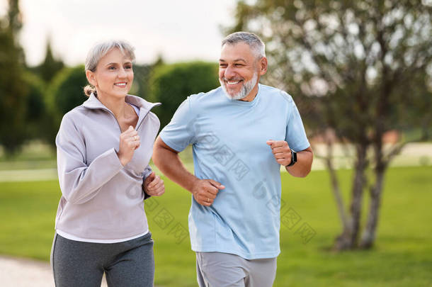 快乐的老夫妇穿着运动服在公园里跑来跑去，可爱的退休人员在阳光灿烂的早晨慢跑，面带微笑地望着对方。健康生活方式概念