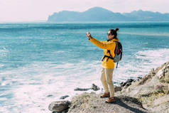 一个背对着大海的带着电话的旅行者一位旅行博客作者在他的手机上，在大海的背景下拍摄了一个照片视频。旅行和积极的生活方式的概念。靠海的游客