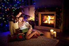 圣诞节前夕，快乐的年轻妈妈和她的小女儿在一个舒适的黑暗客厅里的壁炉边打开了一份神奇的圣诞礼物.
