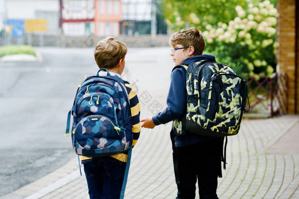 两个背着背包或背包的小男孩。学童在上学的路上。健康的微笑的孩子，兄弟和最好的朋友在外面的街上离开家。回学校去快乐的兄弟姐妹.