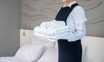 酒店房间服务使房间年轻的女人女服务员持有堆栈折叠白色浴毛巾和站下一个的床上客人房间