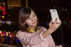 有吸引力的马来西亚女穿耳机而且持有细胞电话与城市灯背景