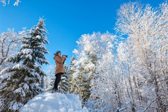 年轻的女人走冬天森林和采取照片在白雪覆盖的树