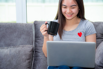 亚洲女人持有咖啡杯微笑和看在工作工作远程与移动PC的沙发的房子可爱的女孩青少年快乐使用互联网电脑沟通从首页女人持有咖啡杯在工作