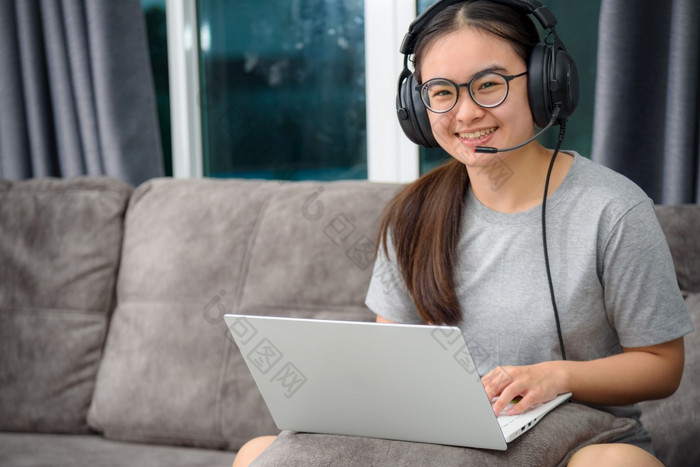 亚洲年轻的女人学生与耳机坐着的沙发微笑看快乐研究在线类大学学习互联网教育十几岁的女孩工作距离移动PC电脑从首页距离学习在线从首页