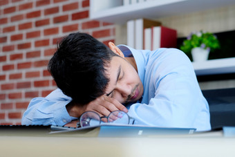 累了亚洲男人。睡觉办公室桌子上年轻的商人与眼镜<strong>过度劳累</strong>和下降了睡着了有创意的休闲男人。睡觉他的工作的地方