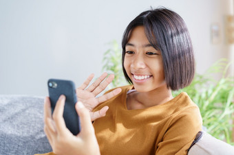 快乐亚洲小女孩视频调用的智能手机和挥舞着问候而坐着的生活房间首页概念放松和生活方式首页