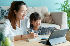 在家教育亚洲家庭与女儿做家庭作业使用平板电脑与妈妈。帮助亚洲妈妈和孩子学习在线与移动PC电脑在一起的生活房间首页