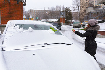 年轻的美丽的女人黑色的外套清洗的雪与折叠绿色长刷从的表面她的车年轻的女人清洗雪从的表面她的车