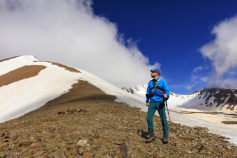 旅游摄影师与狗他的手站的坡白雪覆盖的山凝视它的前和规划爬对的背景明亮的蓝色的和多云的天空旅游摄影师检查的前白雪覆盖的山和准备爬