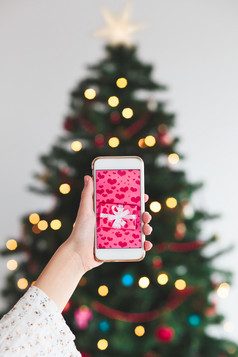 女孩持有的智能手机与包装礼物的屏幕和圣诞节树的背景