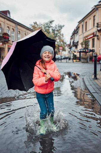 快乐微笑女孩持有大伞跳的水坑在走市中心多雨的悲观的秋天一天小女孩持有大伞跳的水坑多雨的悲观的秋天一天