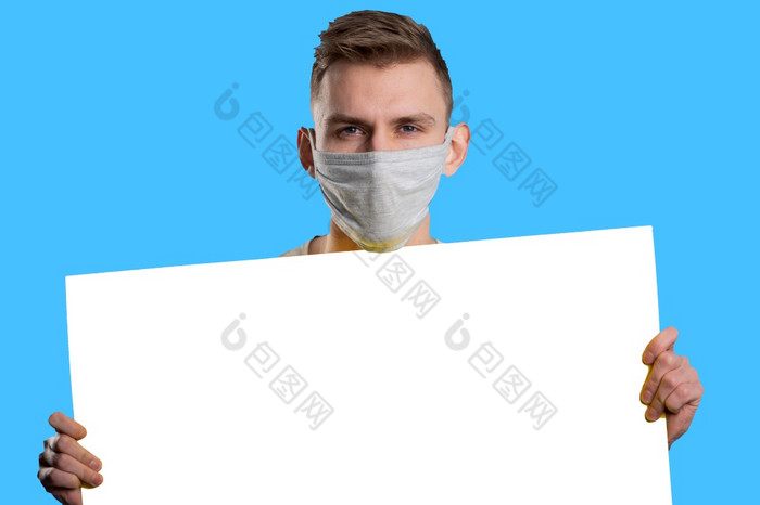 年轻的男人。穿医疗面具显示白色纸海报和看相机蓝色的背景广告概念年轻的男人。穿医疗面具显示白色纸海报和看相机蓝色的背景